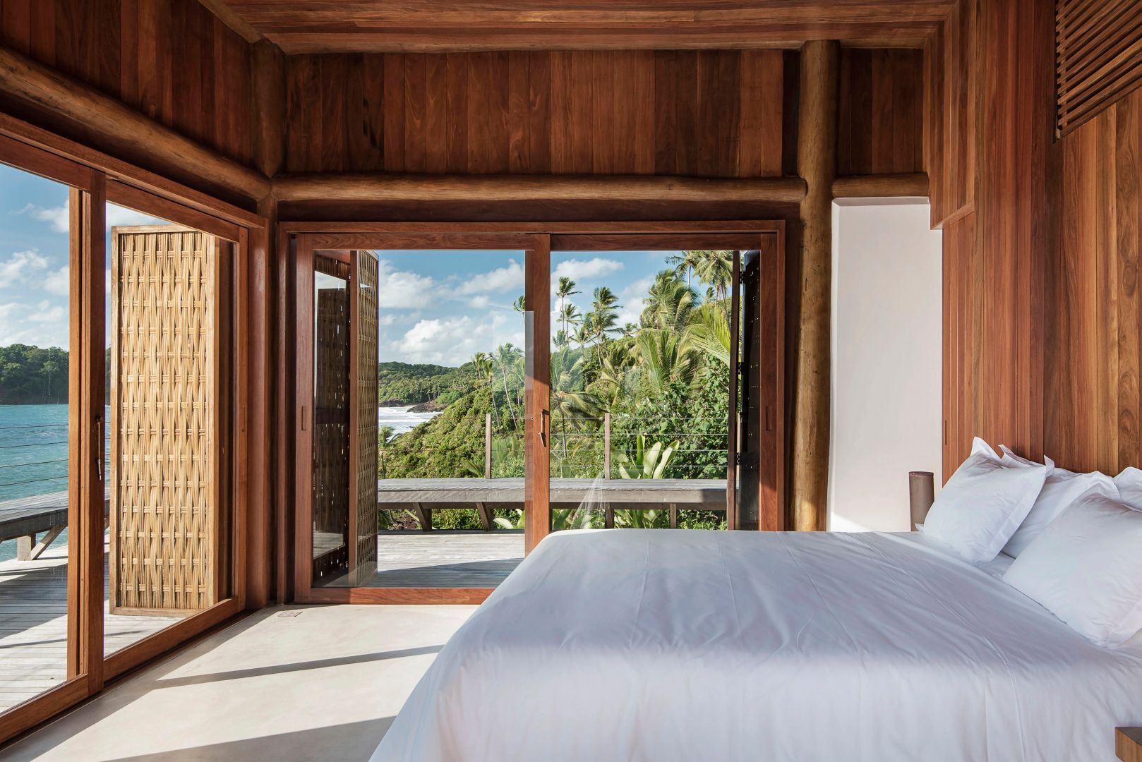Schlafzimmer mit weißem Bett und großer Schiebetür mit Blick auf Tropen und Meer.