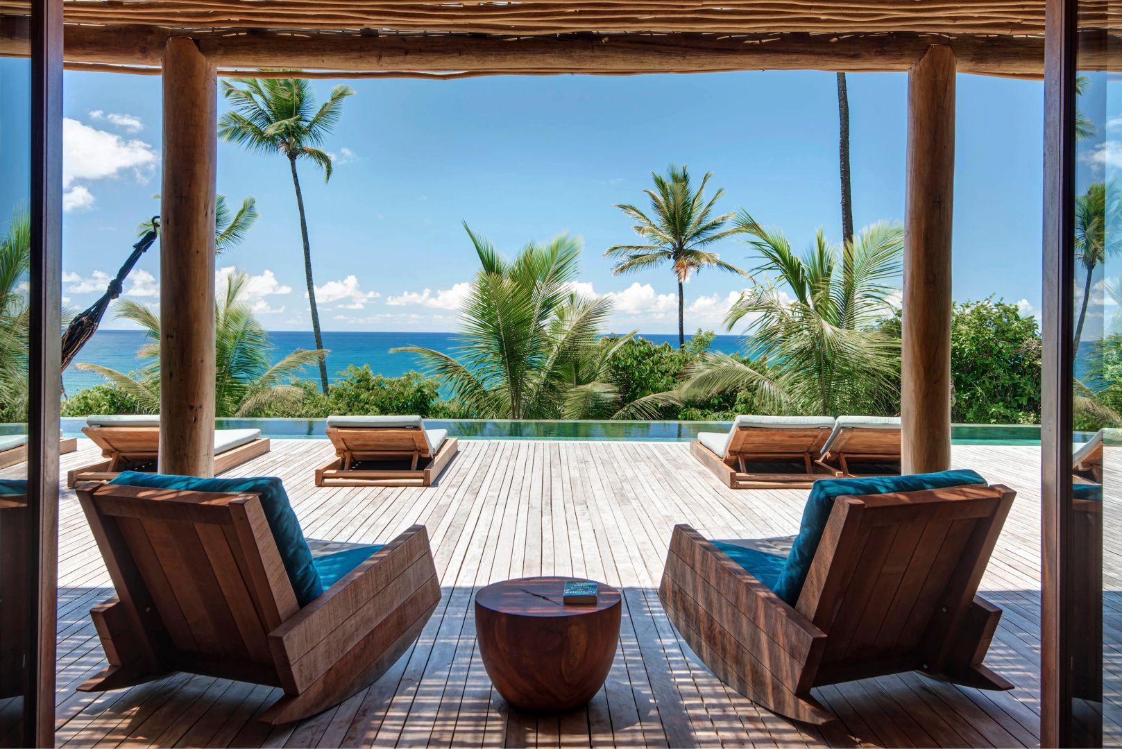 Überdachte Holzterrasse mit Sesseln, Pool und Blick auf Meer und Palmen.