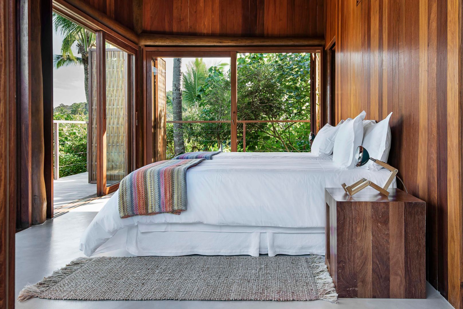 Schlafzimmer mit Holzverkleidung, buntem Bettvorleger und Blick in die Vegetation.