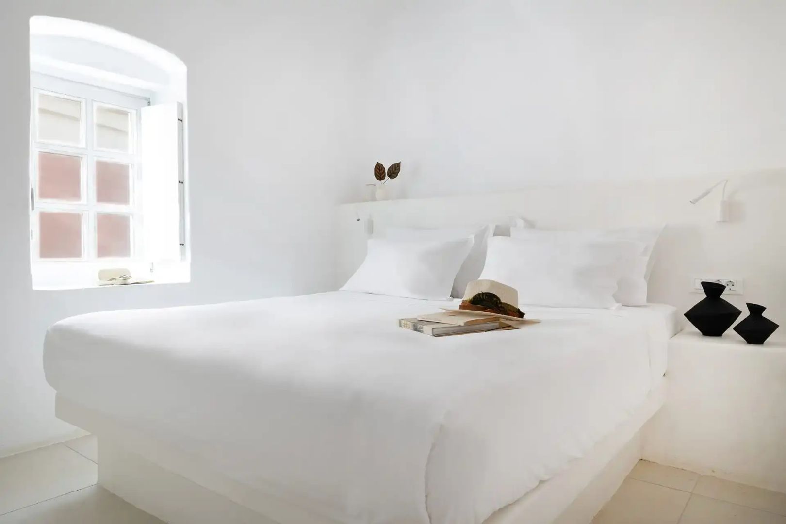 Weiß bezogenes Doppelbett mit Hut und Buch vor einem hellen Fenster
