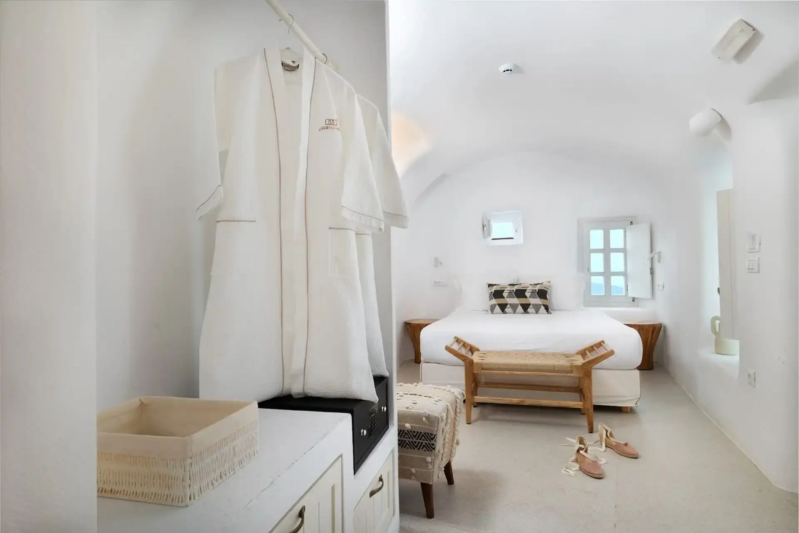 Helles Schlafzimmer mit Doppelbett, Holzbank und aufgehängten weißen Bademänteln.