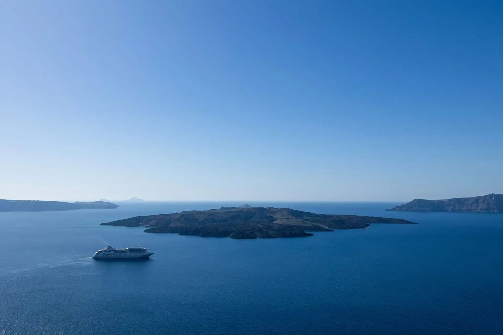 Blick auf eine vulkanische Insel im blau schimmernden Meer unter blauem Himmel mit einem Kreuzfahrtschiff.