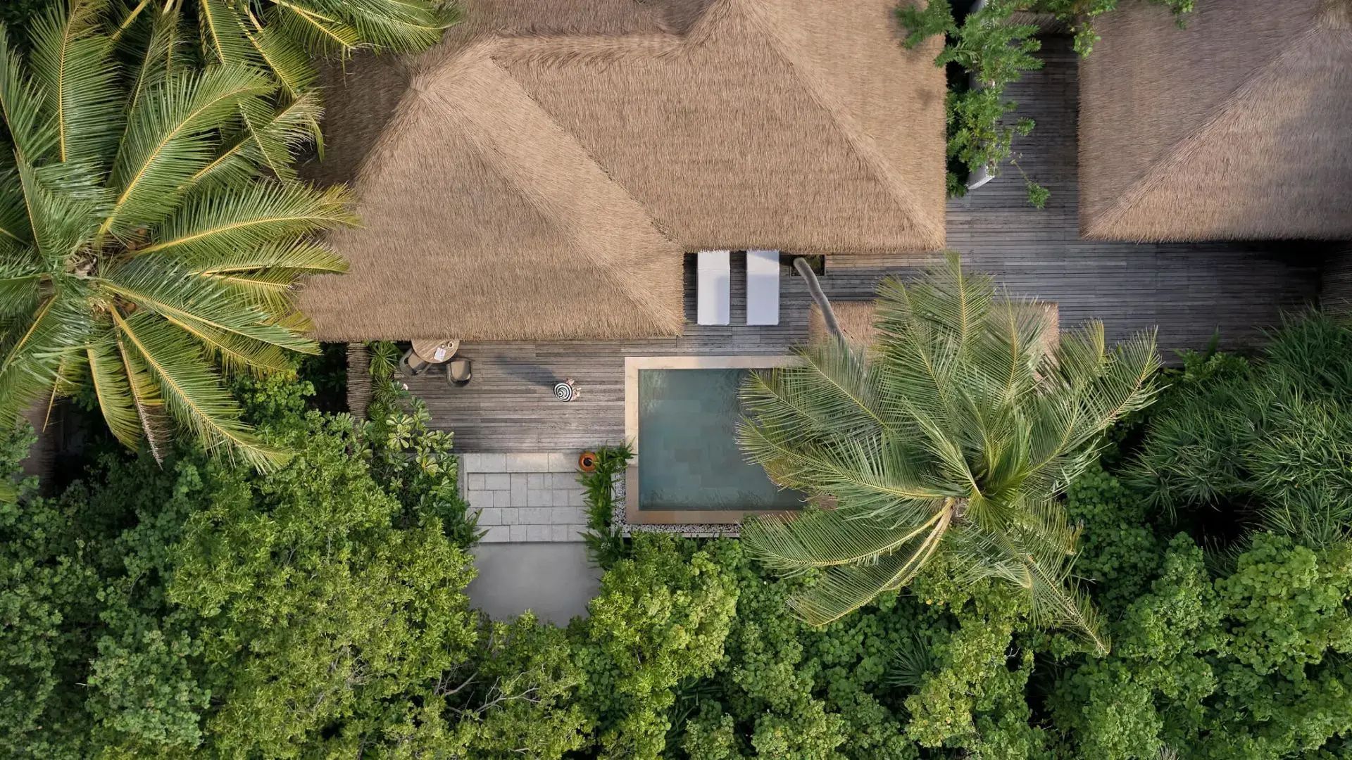 Vogelperspektive auf eine Reetdachvilla mit Holzterrasse, Pool und dichtem Tropengrün.