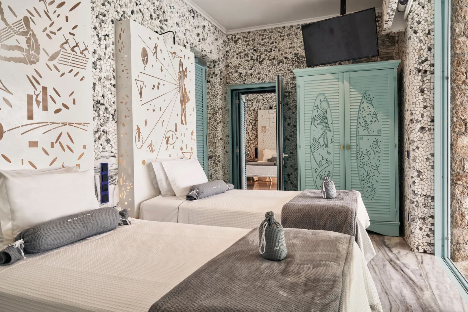 Hotelzimmer mit zwei Einzelbetten, Mosaikwand und türkisblauem Holzschrank.