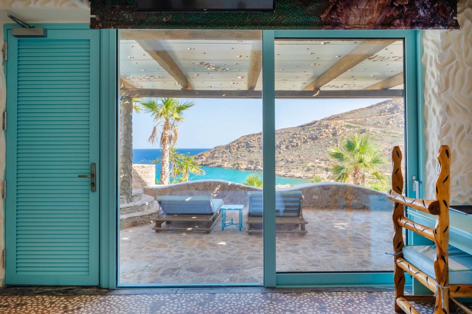 Blick durch ein großes Fenster auf eine Terrasse mit zwei Sonnenliegen und Meerblick.