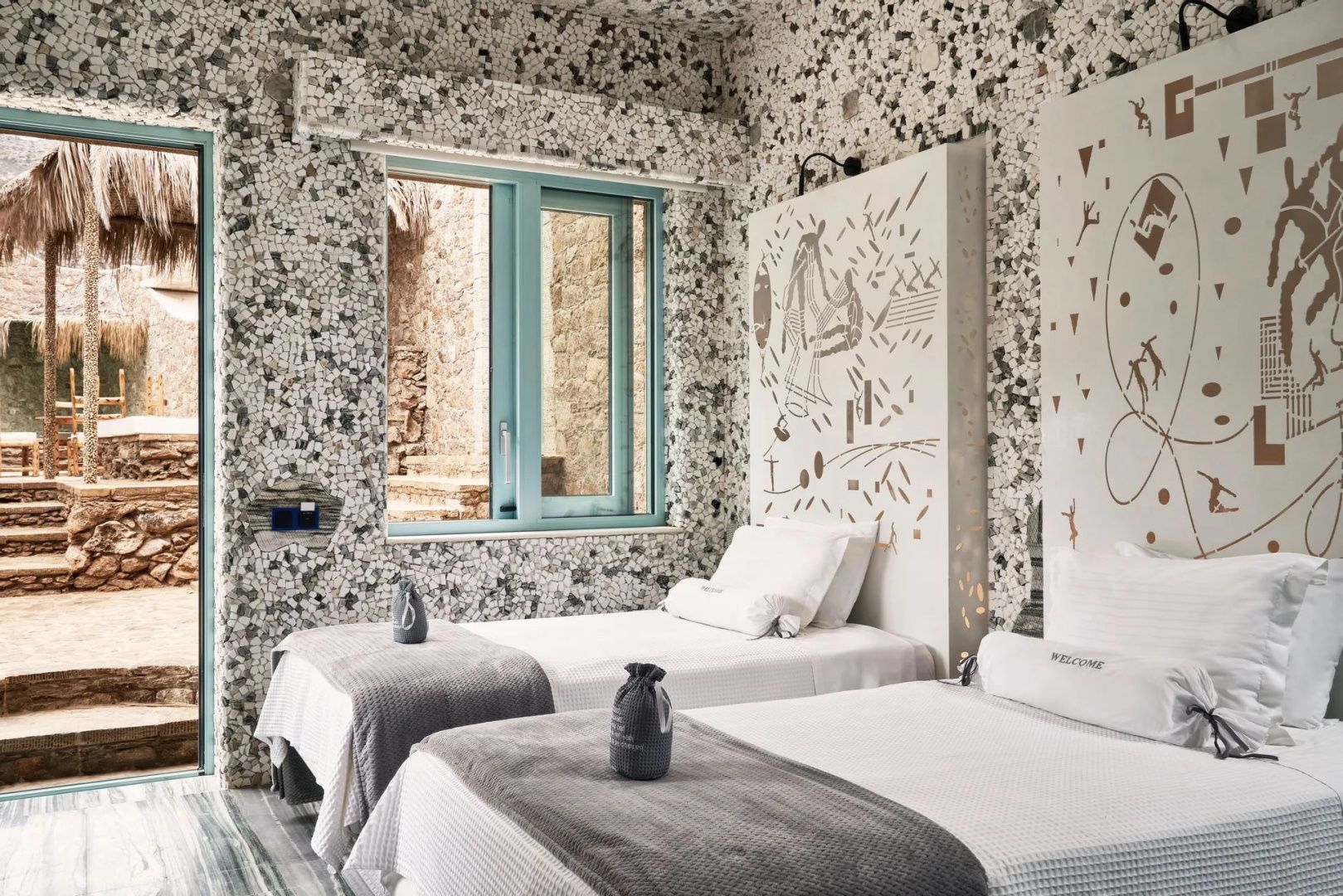 Zweibettzimmer mit Terrazzowänden, weißen Bettdecken und kunstvollen Zeichnungen an den Betten.