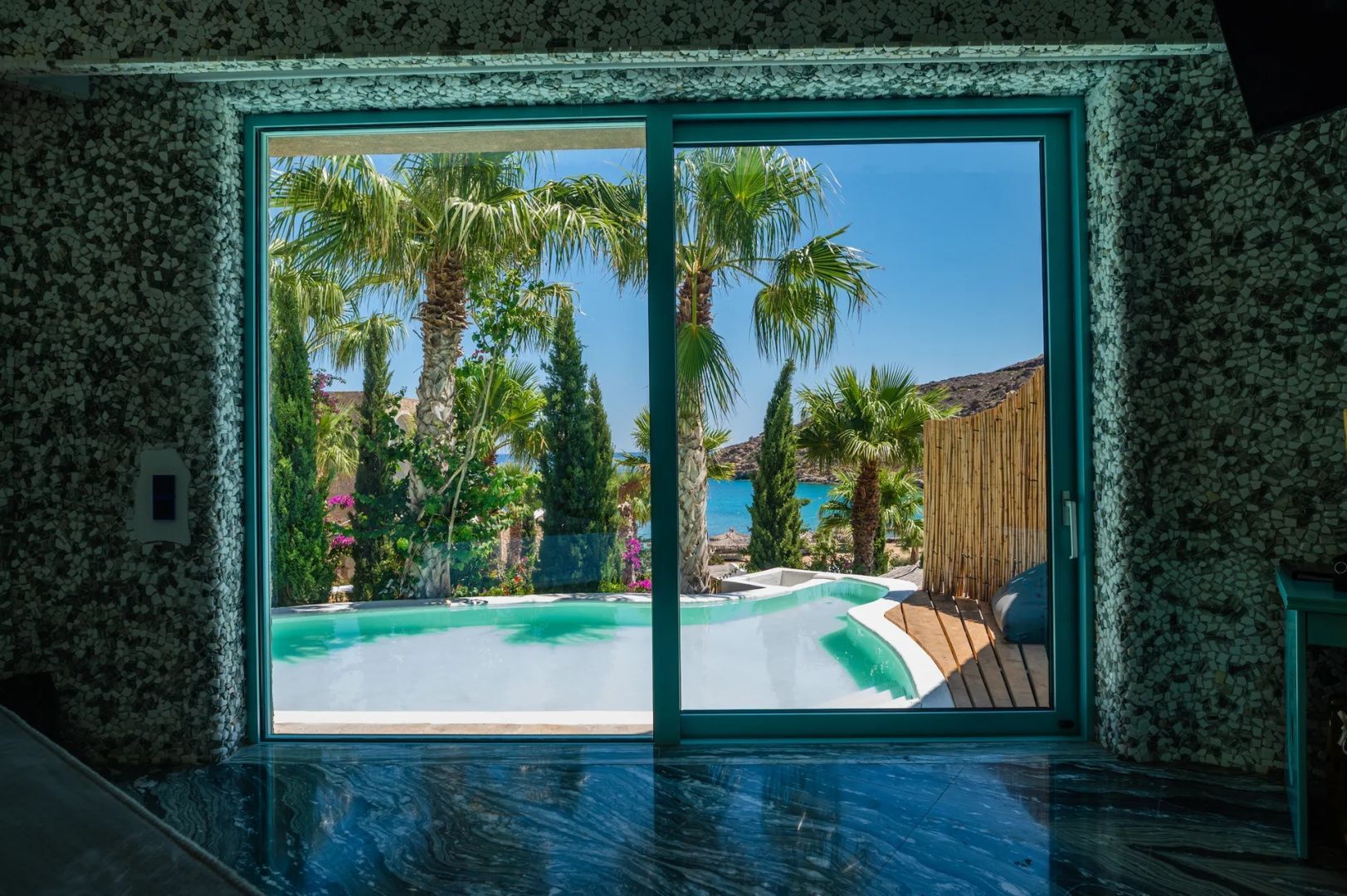Blick durch eine Glastür auf einen privaten Pool, Palmen und das Meer.