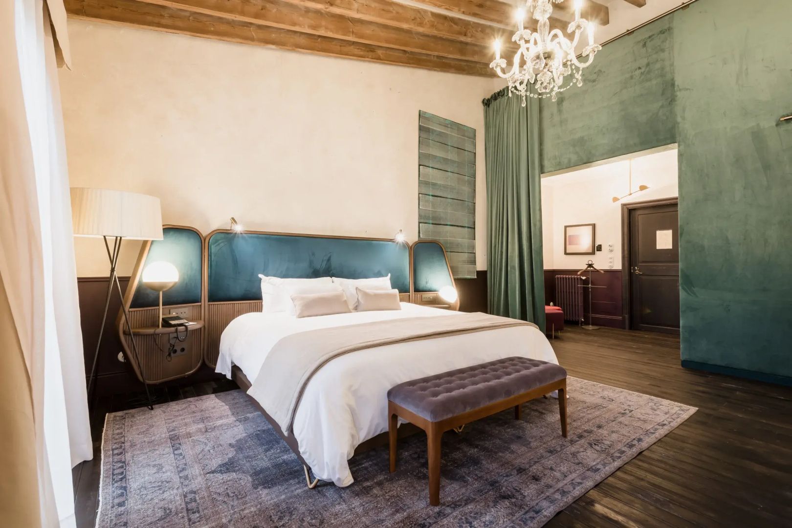 Doppelbett mit blauem Kopfteil, Holzdeckenbalken und Kronleuchter im Hotelzimmer.