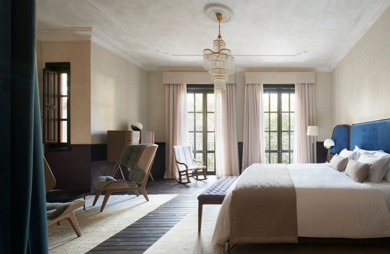 Helles Hotelzimmer mit großem Bett, bequemen Sesseln, Kronleuchter und bodentiefen Fenstern.