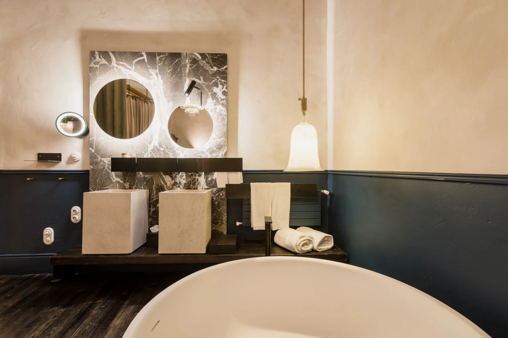 Badezimmer mit Doppelwaschbecken vor einer Marmorwand und runden Spiegeln mit Licht.