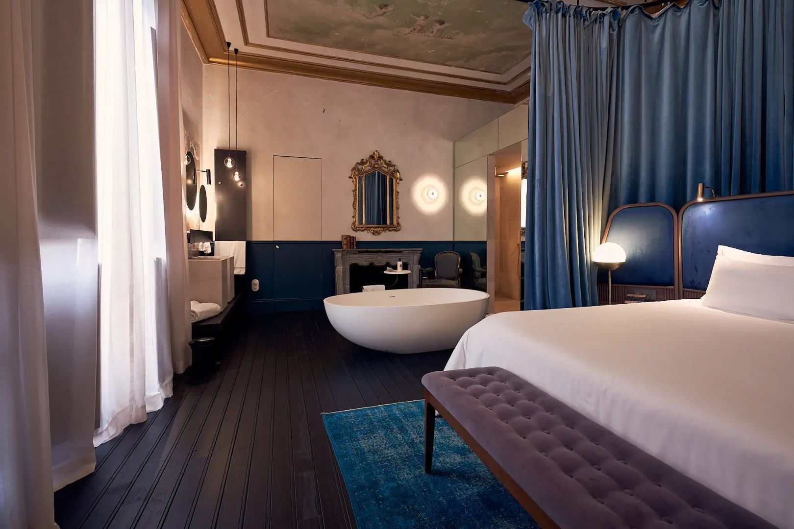 Freistehende weiße Badewanne in einer Suite mit blauem Vorhang und goldenem Spiegel.