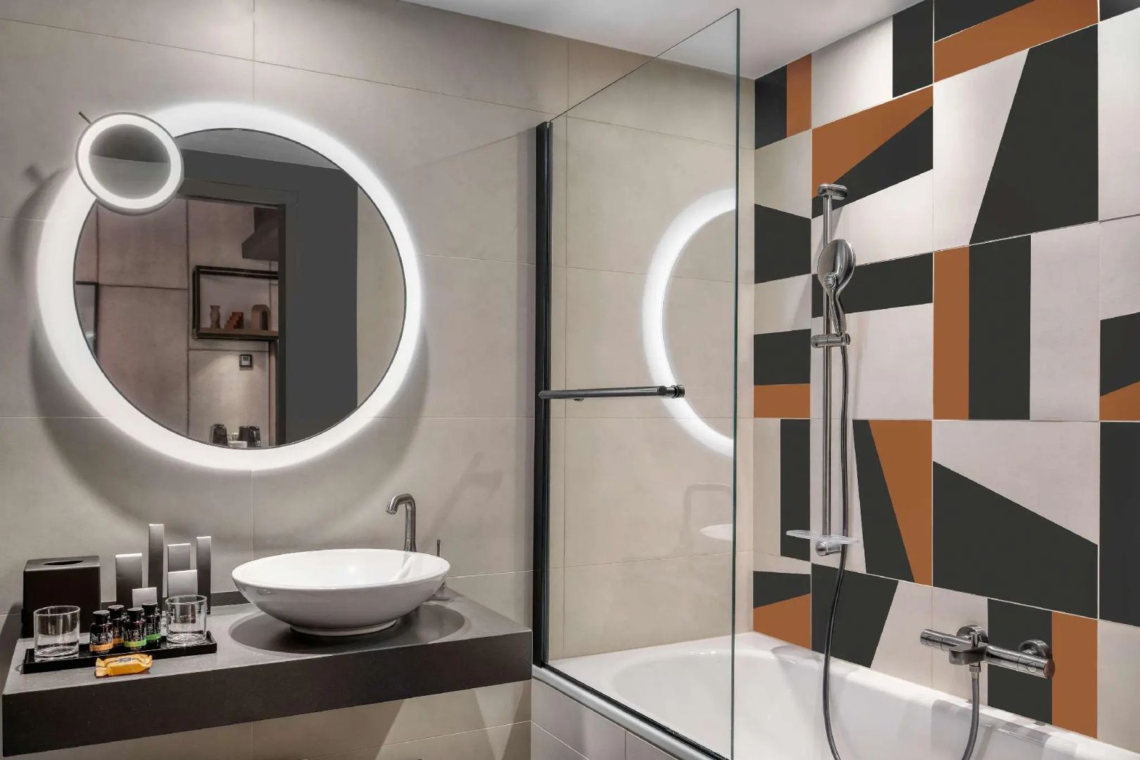 Badezimmer mit beleuchtetem Rundspiegel, Waschtisch und Dusche mit gemusterten Fliesen.