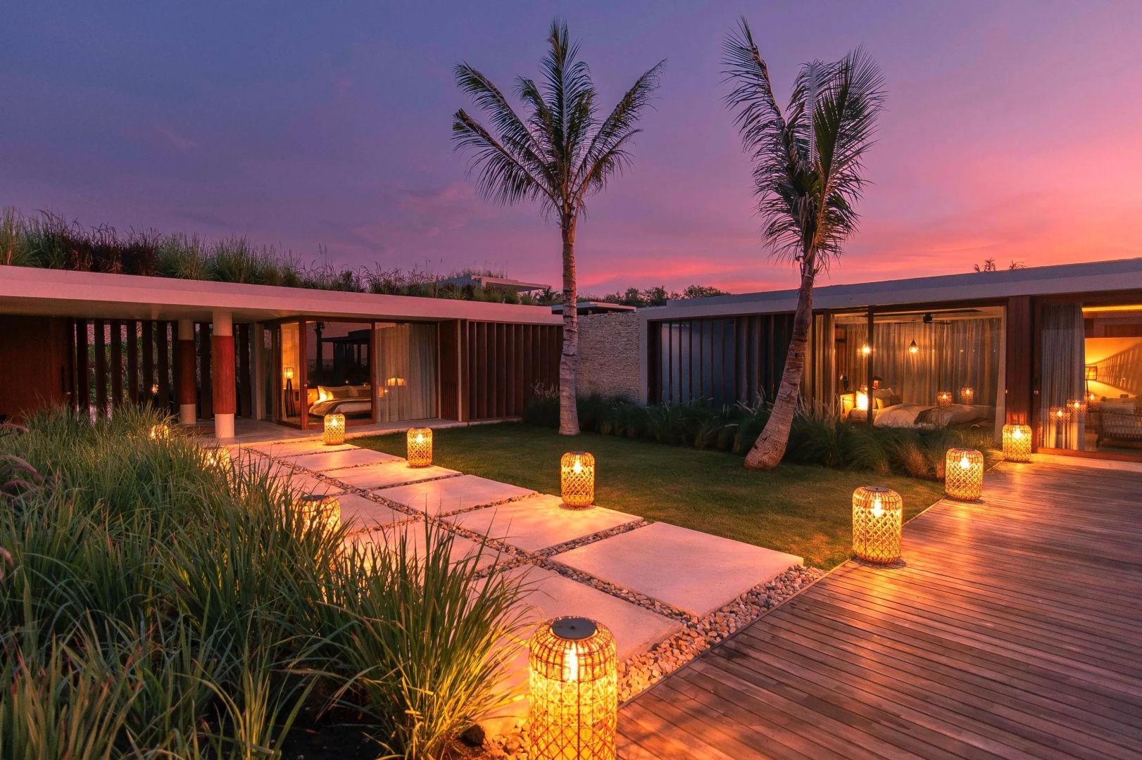 Beleuchteter Gehweg und Palmen vor modernen Bungalows unter rosa Abendhimmel.