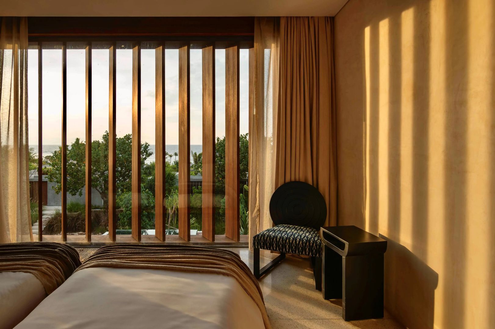 Hotelzimmer mit Holzlamellen am Fenster, Stuhl und warmem Sonnenlicht auf der Wand.