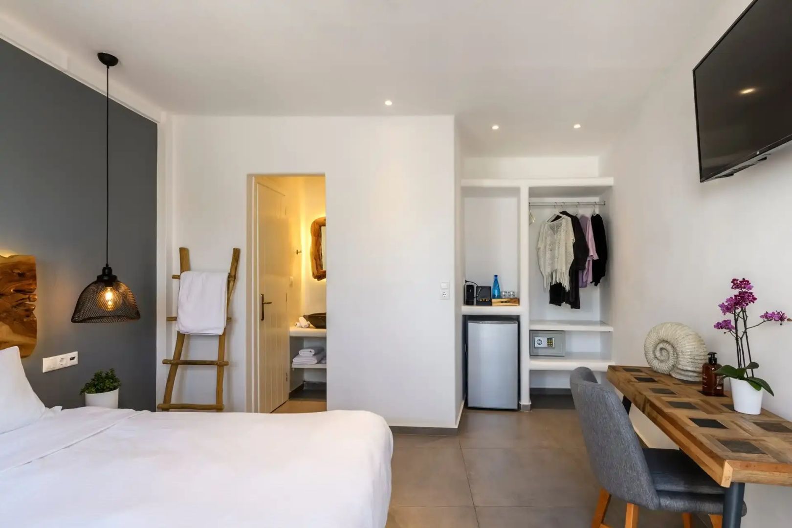 Hell eingerichtetes Hotelzimmer mit Doppelbett, offenem Schrank und Blick ins Badezimmer.
