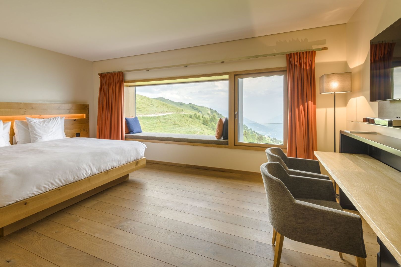 Helles Hotelzimmer mit Holzboden, einem Bett und einem großen Fenster mit Sitznische und Blick auf Berge.
