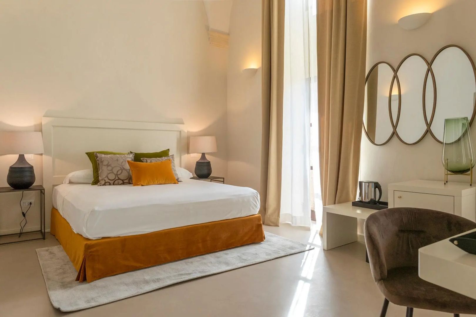 Hell eingerichtetes Hotelzimmer mit großem Doppelbett, hohen Vorhängen und Schreibtisch.