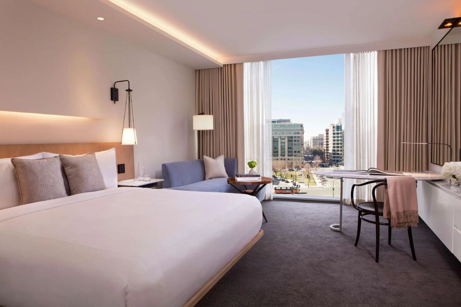 Helles Hotelzimmer mit großem Doppelbett, blauem Sofa und Blick durch die Fensterfront auf die Stadt.