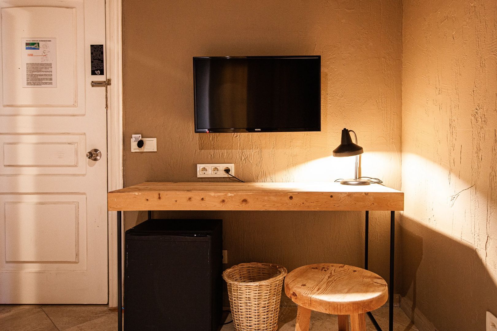 Schreibtisch aus Holz mit Tischlampe, Fernseher an der Wand und Mini-Kühlschrank darunter.