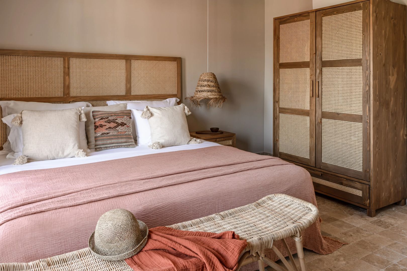 Doppelbett mit rosa Decke, Rattan Kopfteil und Schrank aus Holzgeflecht.