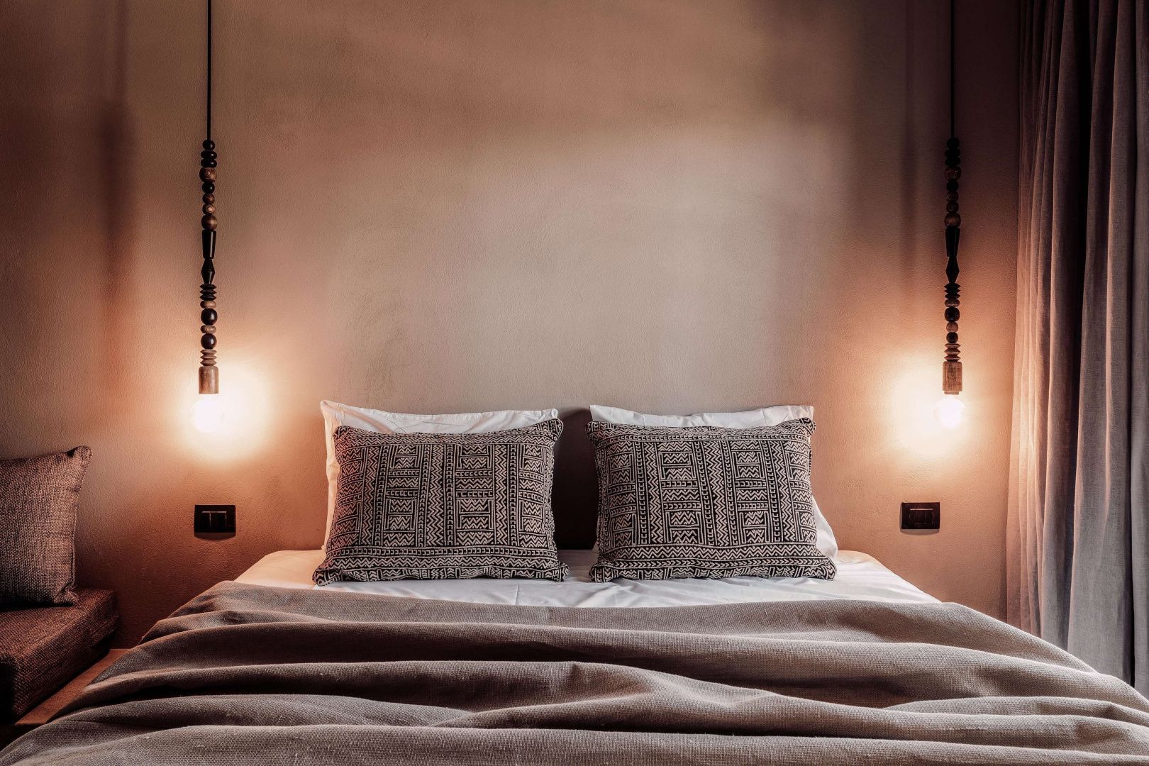 Doppelbett mit gemusterten Kissen und warmem Licht hängender Lampen in erdigen Farbtönen.