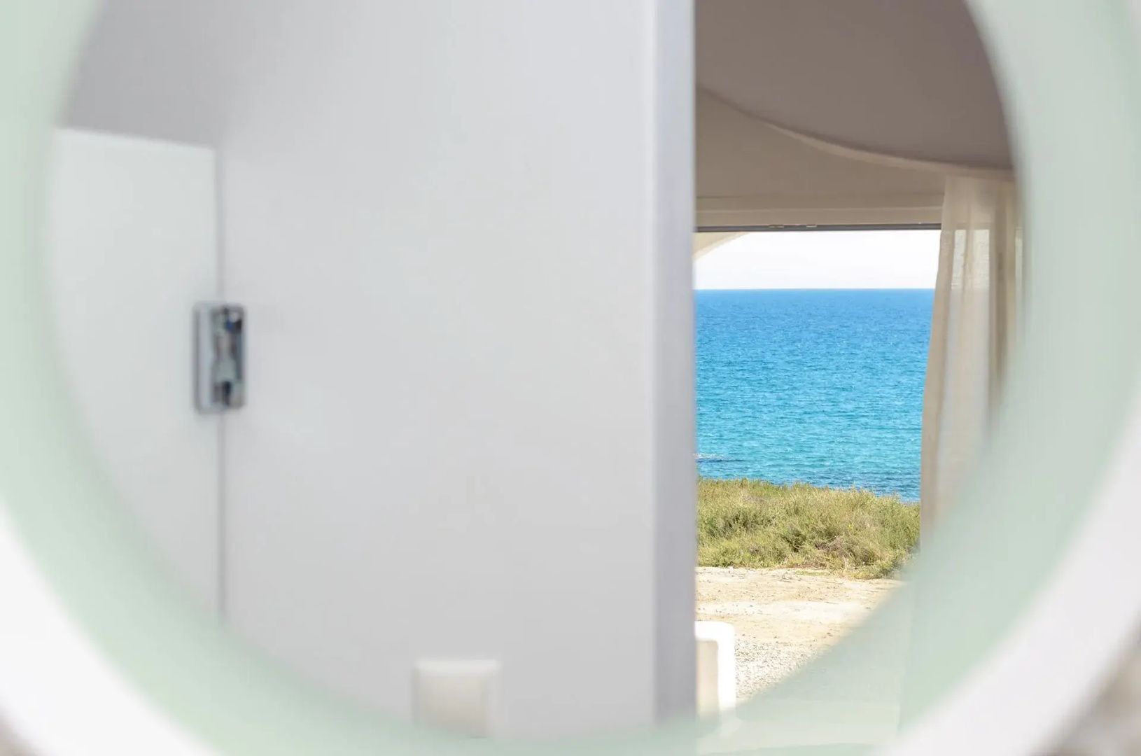 Blick durch einen weißen Durchgang auf das blaue Meer und grüne Küstenlandschaft unter klarem Himmel.