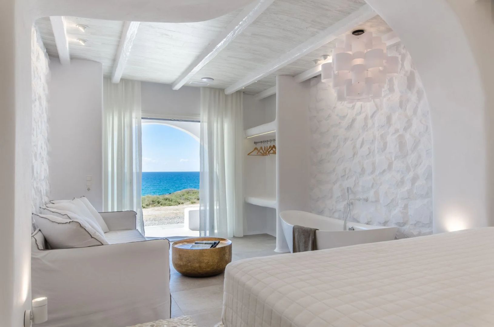 Hell eingerichtetes Zimmer mit Bett, freistehender Badewanne und Blick auf das Meer.