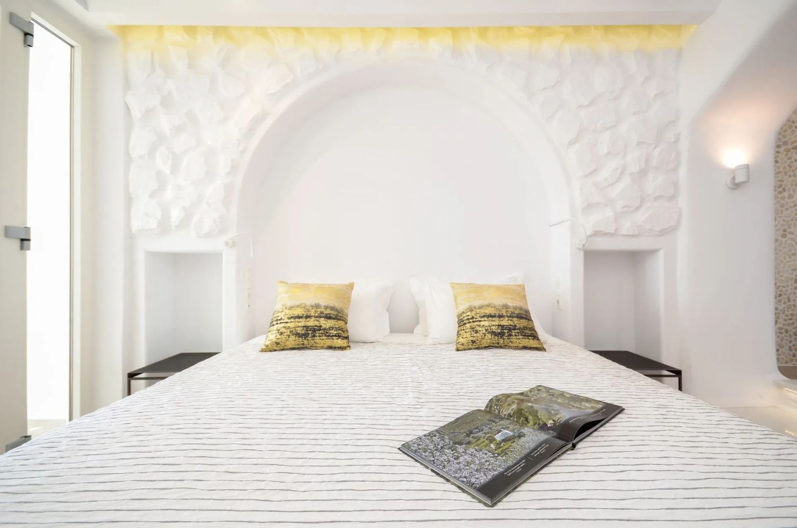 Doppelbett mit hellen Decken, gelben Kissen und aufgeschlagenem Magazin vor weißer Wand.