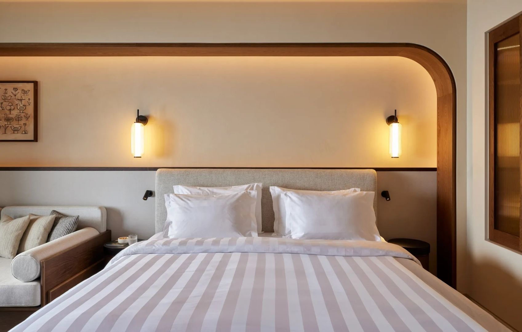 Doppelbett mit gestreifter weißer Bettwäsche und warm beleuchtetem Kopfteil.