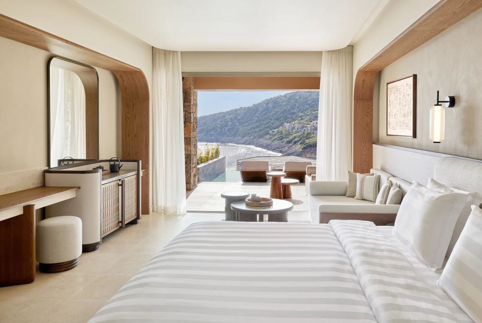 Helles Hotelzimmer mit Sitzbereich und freiem Blick durch die offene Balkontür auf das Meer.
