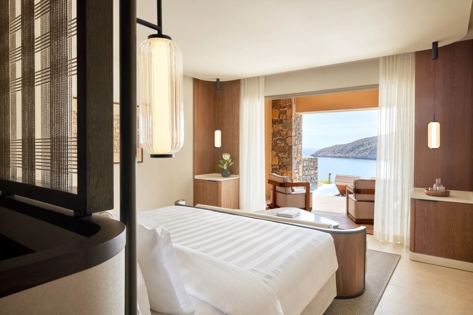 Blick durch das Schlafzimmer mit hängender Leuchte auf die sonnige Terrasse und die Bucht.
