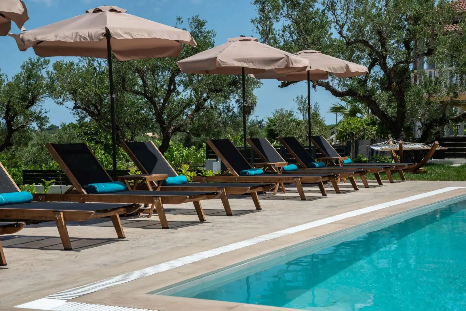 Aufgereihte Holzliegestühle mit Sonnenschirmen stehen am Ufer eines Swimmingpools.