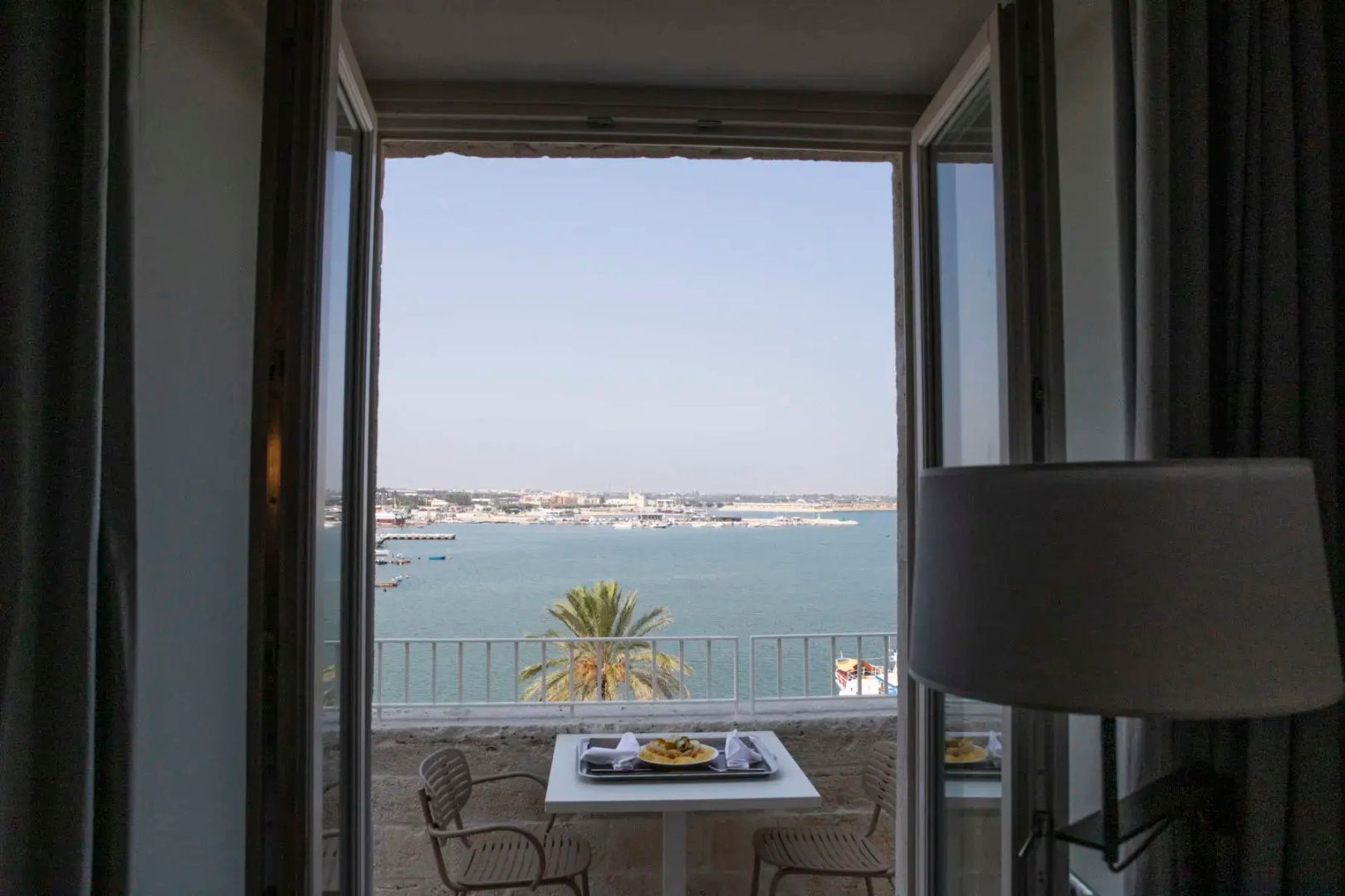 Blick durch zwei geöffnete Fensterflügel auf eine kleine Terrasse und den Hafen am Meer.