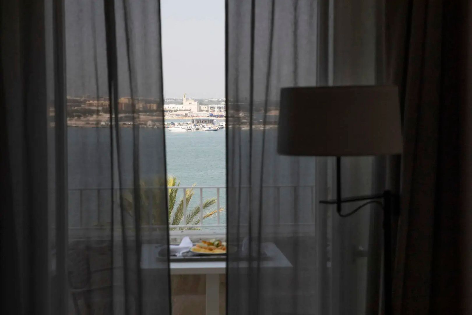 Blick durch dunkle Vorhänge auf einen Balkon mit Aussicht auf Hafen und Meer.