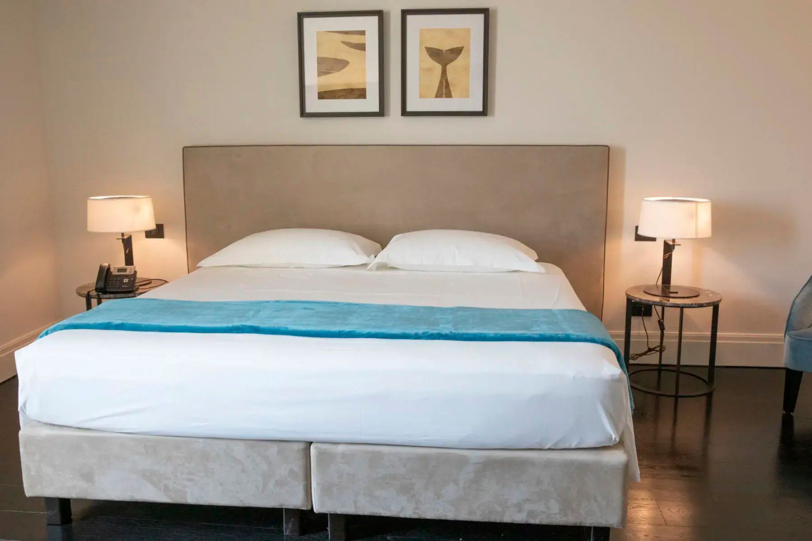 Doppelbett mit beigem Kopfteil, weißer Bettwäsche, blauer Tagesdecke und zwei Bildern an der Wand.