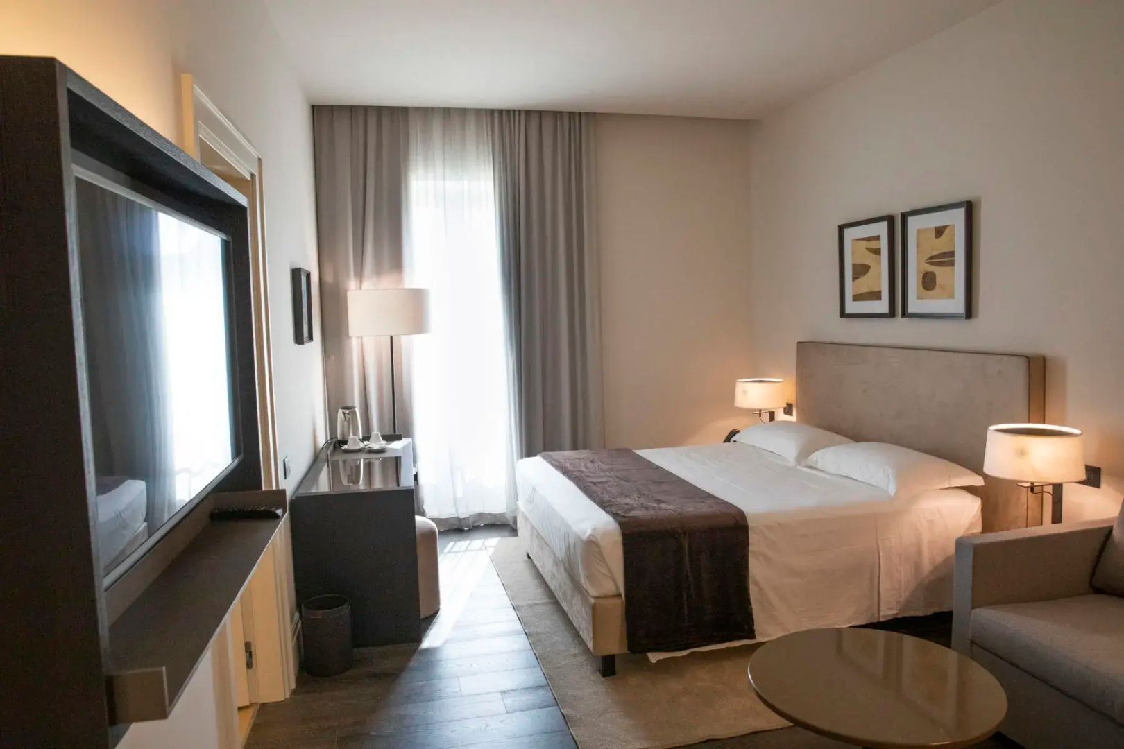 Helles Hotelzimmer mit Doppelbett, Nachttischlampen und Schreibtisch am Fenster.
