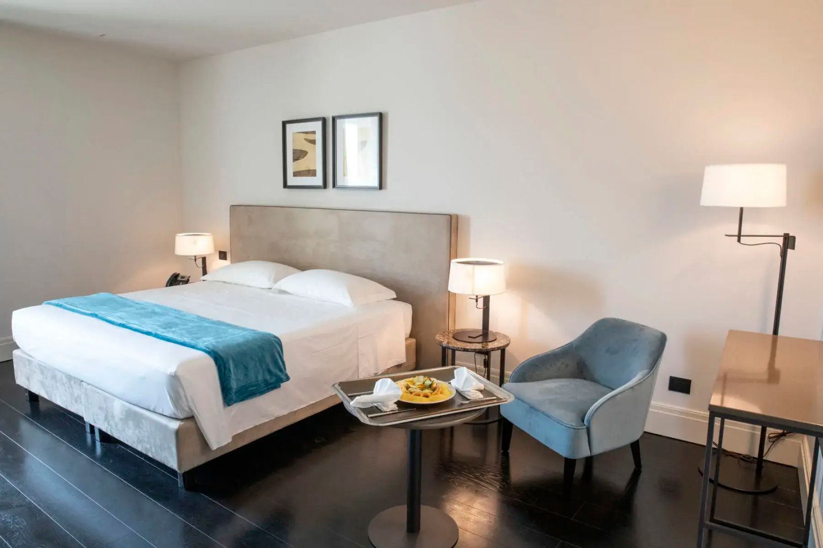 Hotelzimmer mit großem Doppelbett, blauem Polstersessel und einem Tisch mit Tablett.