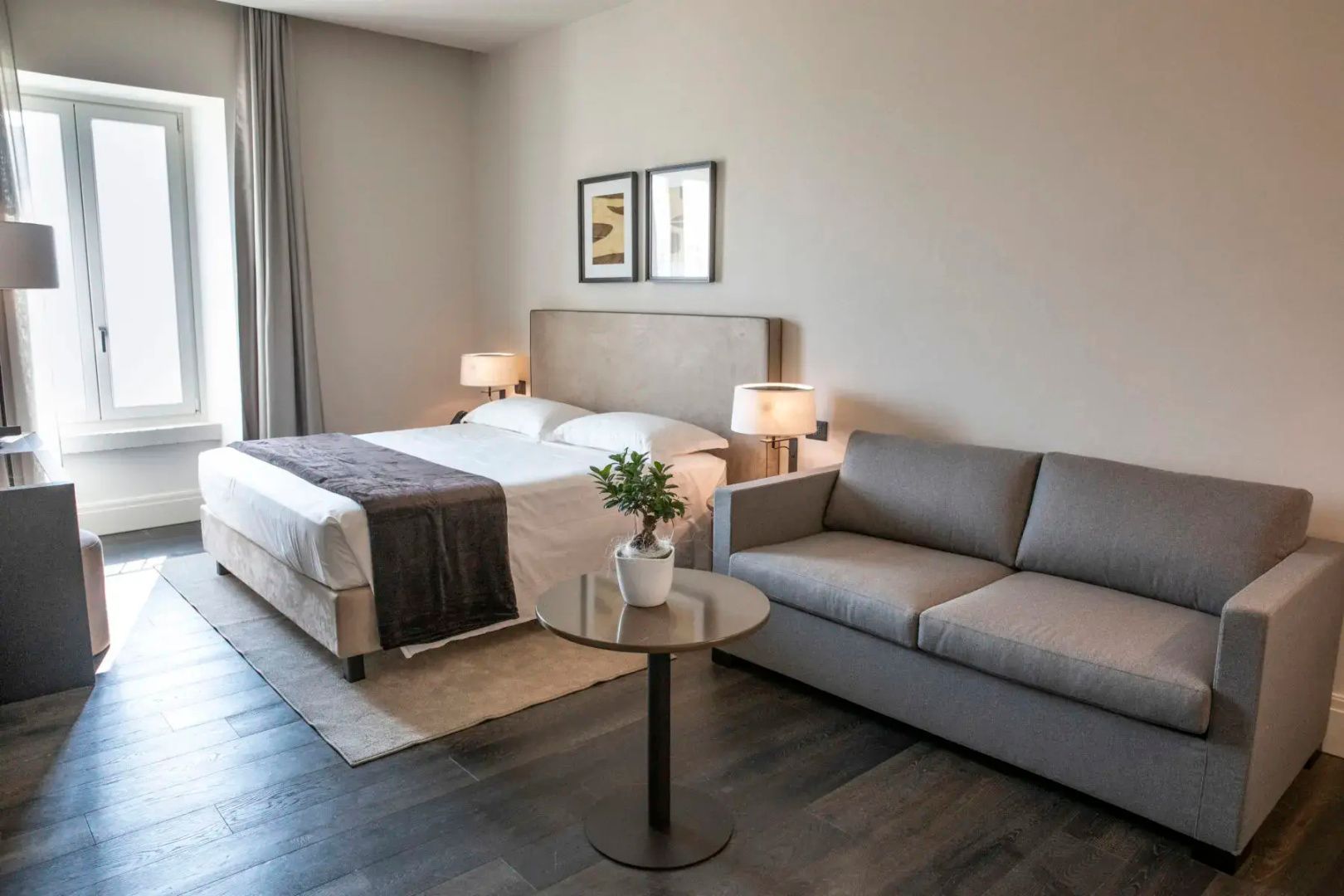 Hotelzimmer mit grauem Sofa, kleinem runden Tisch und Doppelbett im Hintergrund.