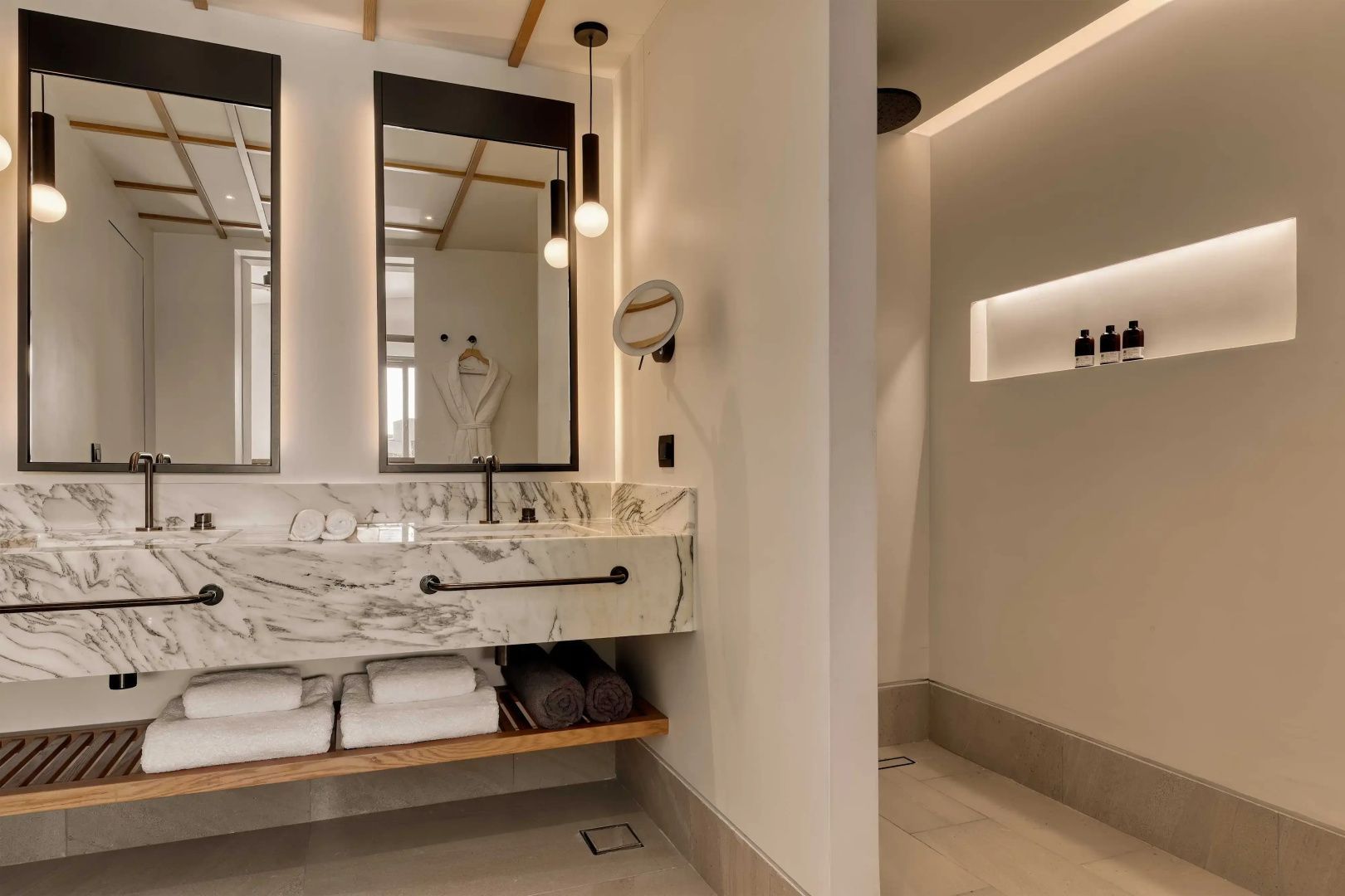 Badezimmer mit Doppelwaschtisch aus Marmor, beleuchteten Spiegeln und Dusche.