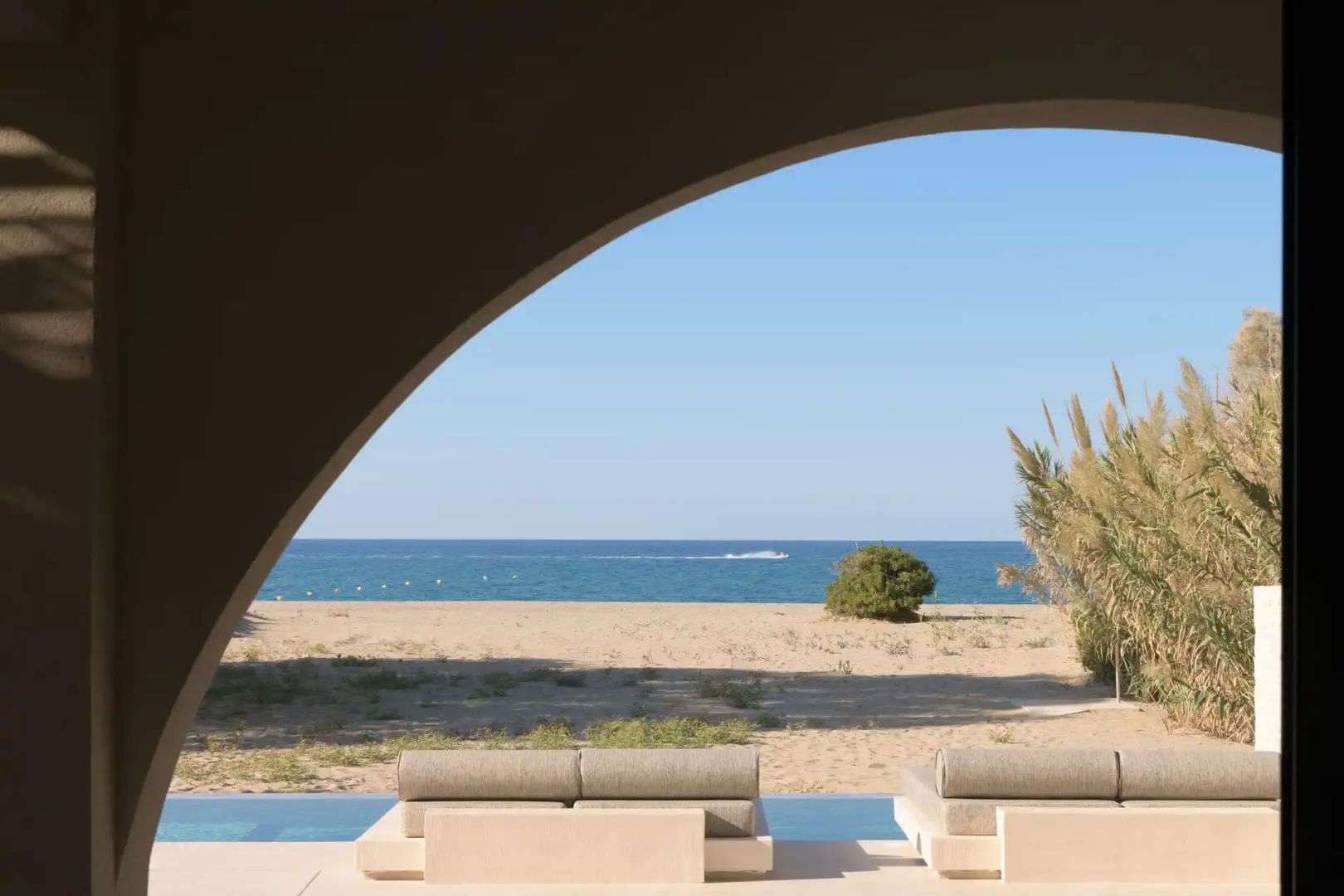 Blick durch einen Rundbogen auf zwei Liegen am Pool, den Sandstrand und das weite Meer.