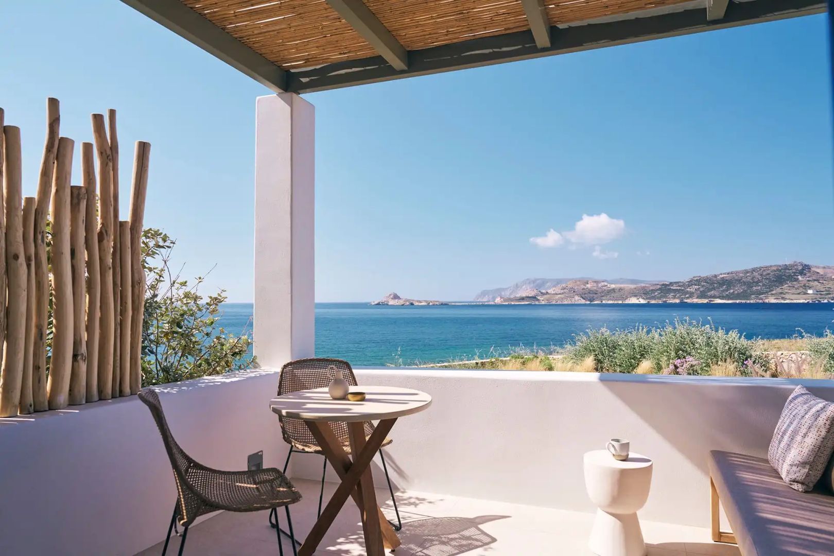 Sonnige Terrasse mit zwei Stühlen, einem runden Tisch und Panoramablick auf das Meer unter blauem Himmel.