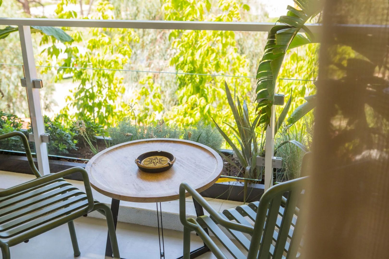 Balkon mit rundem Holztisch, zwei grünen Stühlen und üppiger Bepflanzung im Hintergrund.