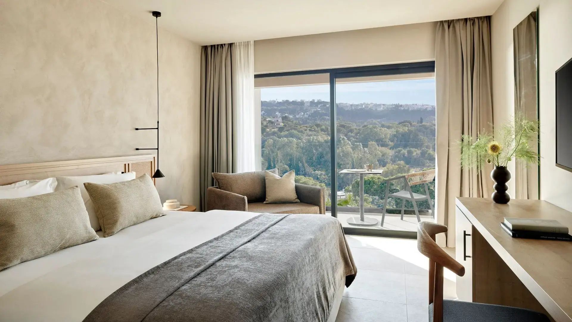 Helles Hotelzimmer mit Doppelbett, Sessel und Balkon mit Blick auf grüne Hügel.