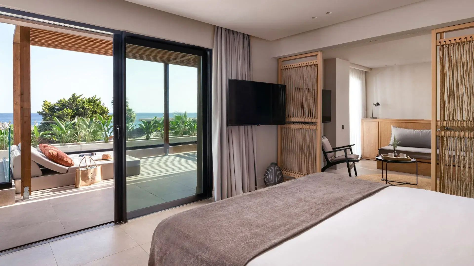 Schlafbereich im Hotelzimmer mit Flachbildfernseher und Schiebetür zur Terrasse mit Meerblick.