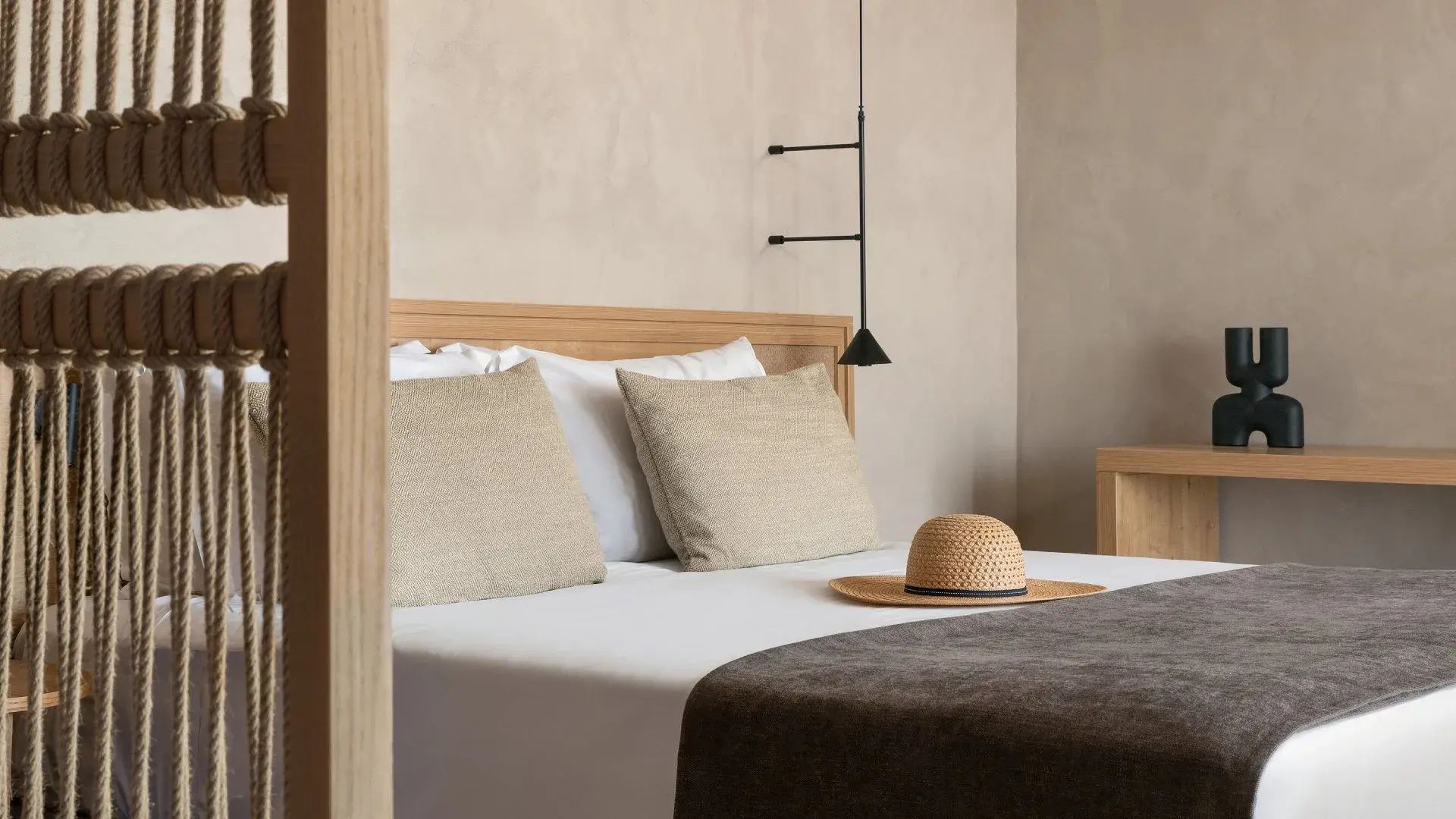 Doppelbett mit Holzkopfteil, hellen Kissen, Strohhut und einer schwarzen Hängelampe im Zimmer.