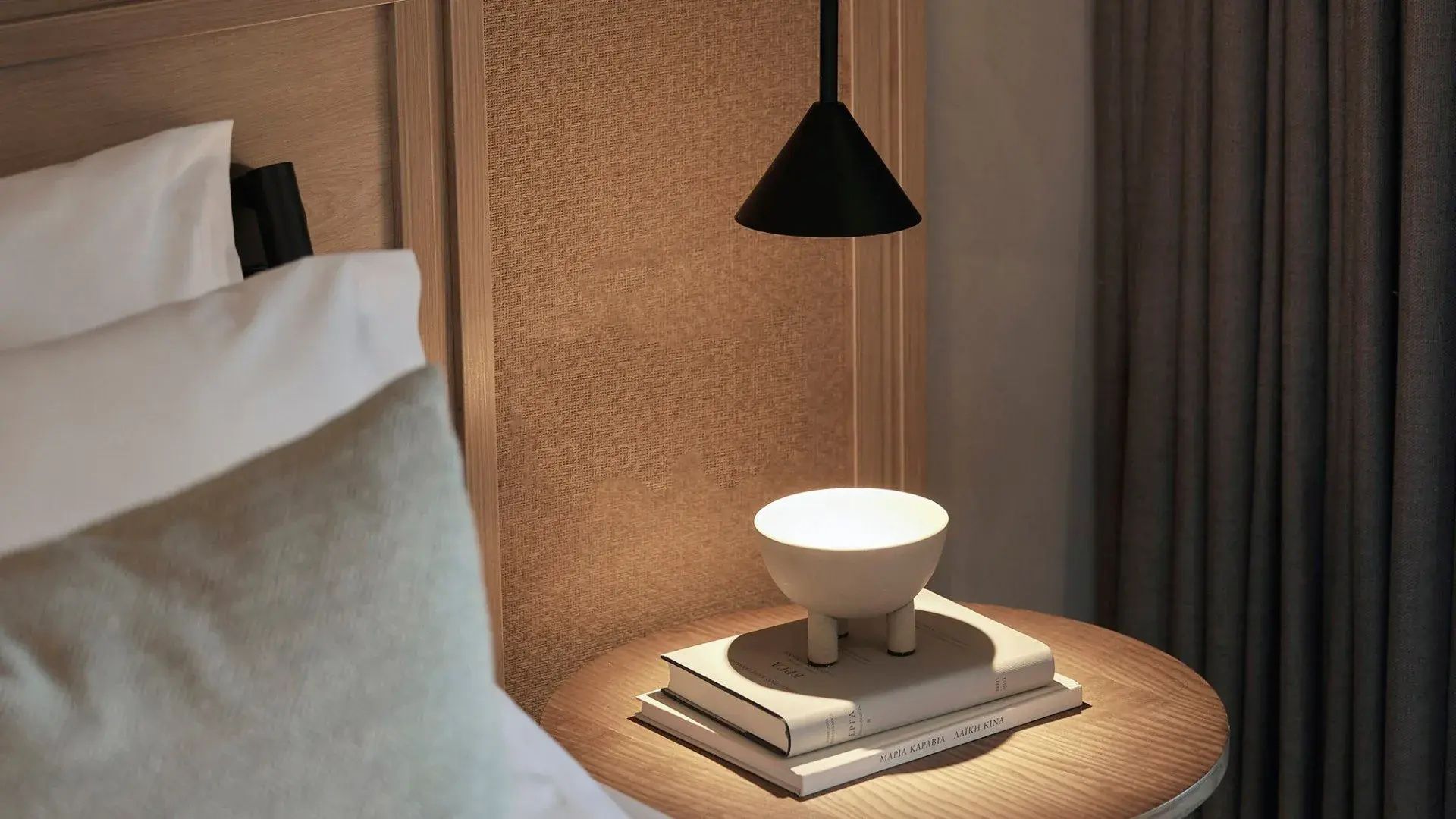 Nachttisch aus Holz mit Büchern, einer Tischlampe und schwarzer Hängeleuchte vor dem Kopfteil des Bettes.
