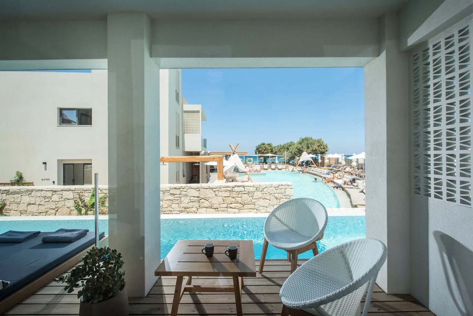 Blick von einer überdachten Terrasse mit Stühlen auf einen Pool und das Meer.