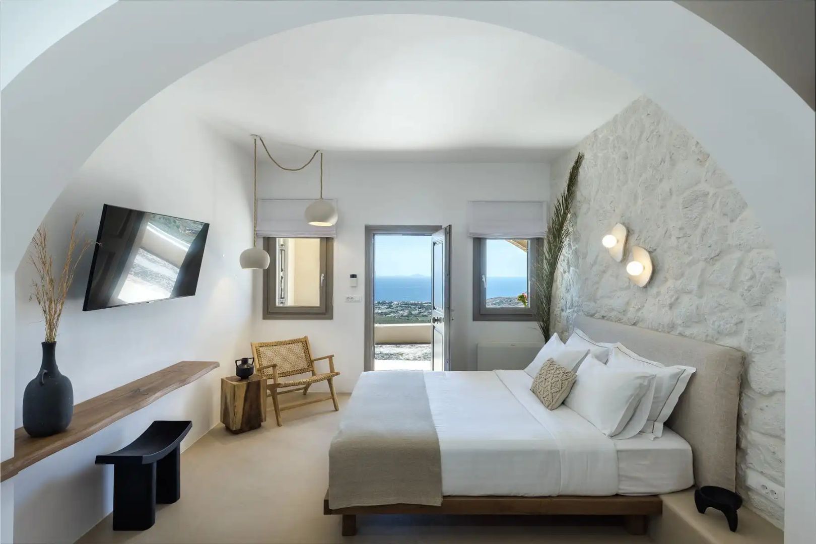 Helles Schlafzimmer mit Holzbett, gewölbter Decke und Meerblick durch die geöffnete Tür.