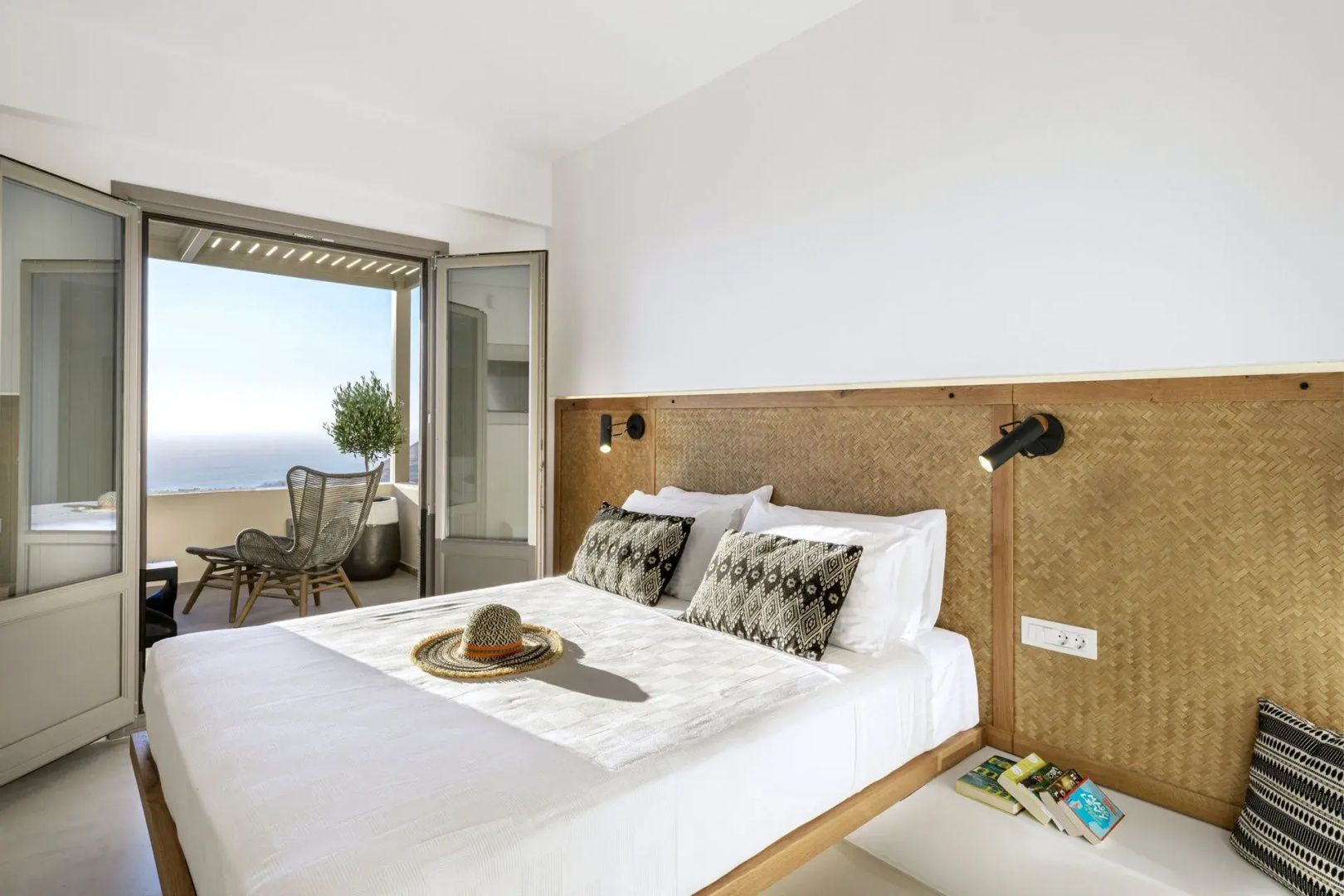 Schlafzimmer mit Kopfteil aus Holz, Strohhut auf dem Bett und Tür zur Terrasse mit Meerblick.