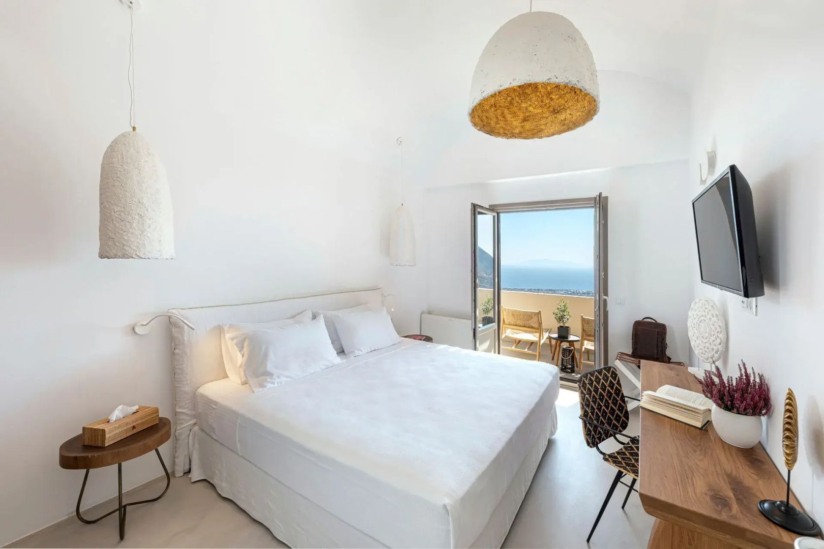 Helles Schlafzimmer mit weißem Doppelbett, Schreibtisch und geöffneter Balkontür mit Meerblick.